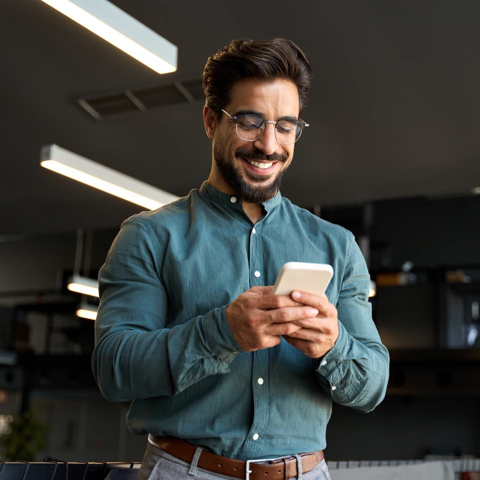 man in button up shirt standing and smiling while looking at his cellphone in his hands
