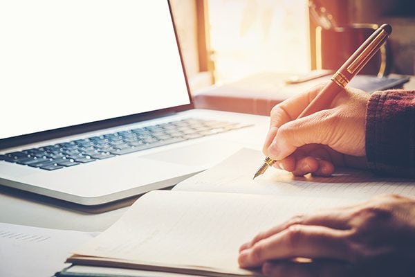 person writing in a notebook in front of a laptop on a table