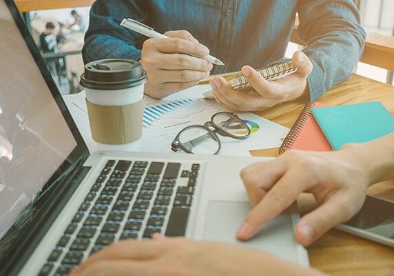 two people working at a table one on a laptop and the other writing on a notepad