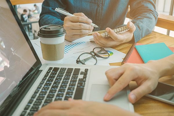 two people working at a table one on a laptop and the other writing on a notepad