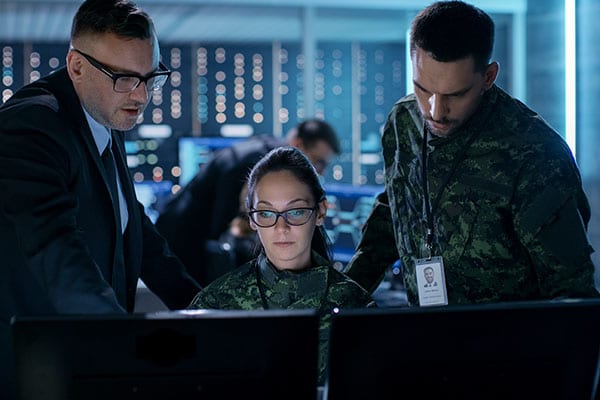 a business professional and a military person looking over the shoulder of another military service member's shoulder at her two monitors in discussion