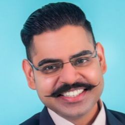 Aditya Sisodia PCC headshot photo, man with dark hair and mustache and glasses smiling light blue background