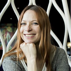 Vibeke Foss headshot photo, smiling woman with red hair and fist under chin in front of white sculpture wall