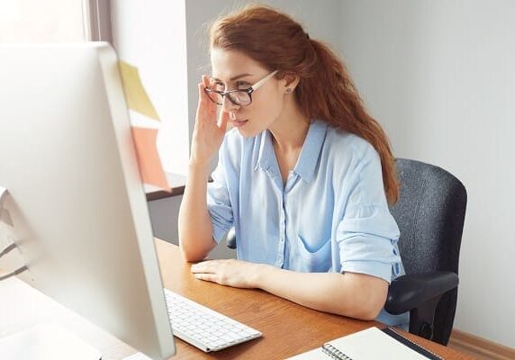 woman with glasses on sitting at a desk looking at a website on a computer monitor